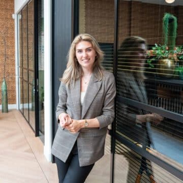 Glimlachende vrouwelijke adviseur leunt tegen een glazen wand in een licht, modern kantoor met planten.
