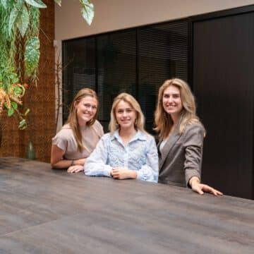 Drie glimlachende vrouwelijke medewerkers achter een tafel in een modern kantoor met planten en bloemen.