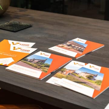 Vier oranje brochures van Vind Makelaardij liggen uitgespreid op een donkere houten tafel.