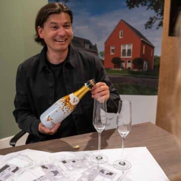 Adviseur opent lachend champagne aan tafel met plattegronden bij een afbeelding van een nieuwbouwhuis.