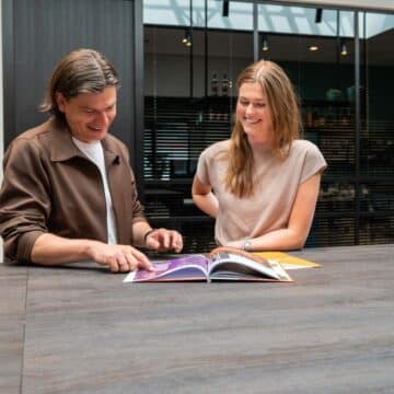 Twee medewerkers bekijken samen lachend een brochure aan een tafel in een modern kantoor.