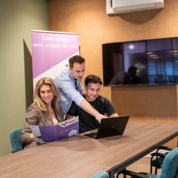 Drie medewerkers bekijken samen een laptop en brochure aan een vergadertafel in een modern kantoor.