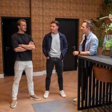 Drie mannen in gesprek in een modern kantoor met houten wandpanelen en decoratieve planten.