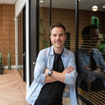 Lachende mannelijke adviseur leunt met gekruiste armen tegen een glazen wand in een modern kantoor.