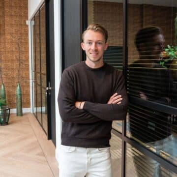 Jonge mannelijke adviseur leunt met gekruiste armen tegen een glazen wand in een modern kantoor.