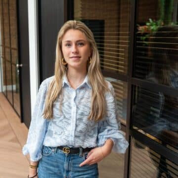 Een jonge vrouwelijke adviseur staat glimlachend in een moderne kantooromgeving.