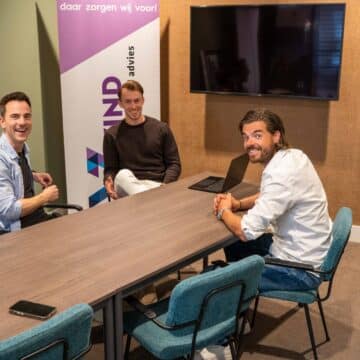 Drie lachende mannelijke medewerkers aan een vergadertafel in een moderne vergaderruimte.