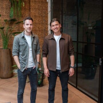 Twee glimlachende mannelijke adviseurs in een modern kantoor met houten vloer en groene plant.