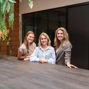 Drie glimlachende vrouwelijke medewerkers achter een tafel in een modern kantoor met planten en bloemen.
