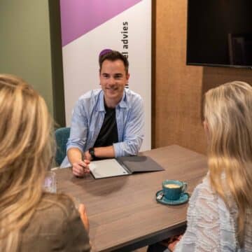 Een adviseur in gesprek met twee klanten aan een tafel, met op de achtergrond een banner van het kantoor.