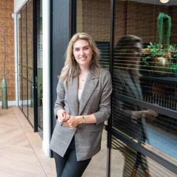 Glimlachende vrouwelijke adviseur leunt tegen een glazen wand in een licht, modern kantoor met planten.