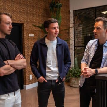 Drie mannen in gesprek in een modern kantoor met houten wanden en planten op de achtergrond.